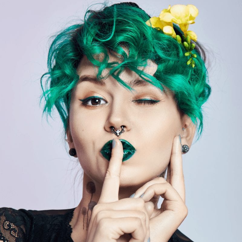 Vrouw met groen kort krullend haar brengt haar vinger naar haar lippen in een shush beweging