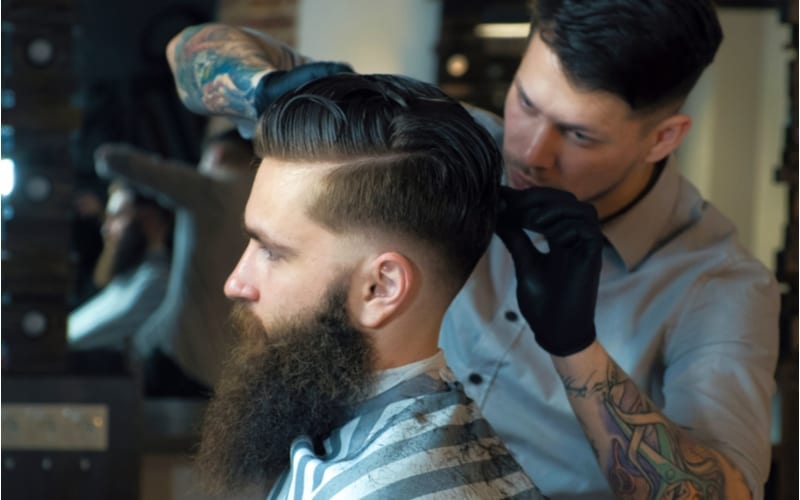 Een man met een korte zijden lang topkapsel zit door een knappe man met baard naar voren te kijken terwijl hij zijn haar laat knippen door de kapper in de kapperszaak