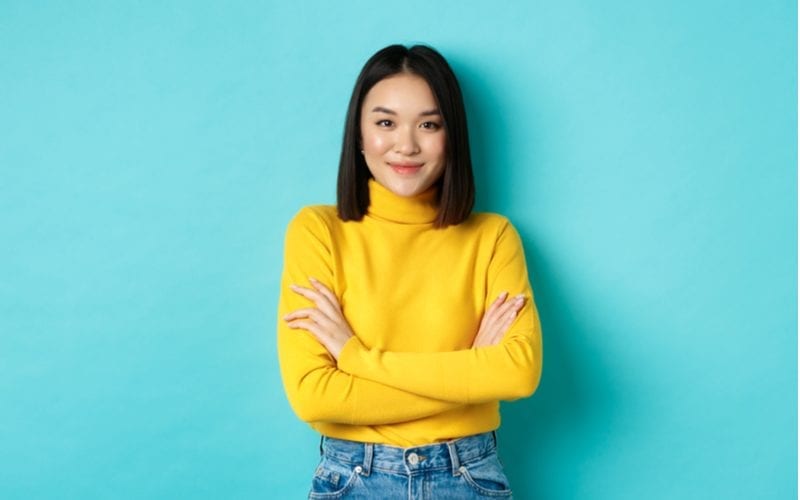 Aziatische vrouw in een gele trui staat haar armen te kruisen en kijkt naar de camera in een blauwe kamer