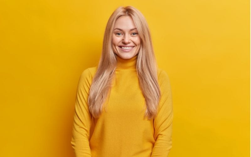 Langharige blonde vrouw staat in een oranje shirt in een oranje kamer met een vierkant gezicht om je te helpen beslissen welk kapsel je moet krijgen