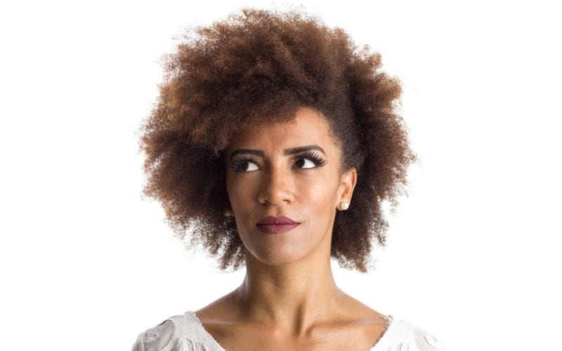 Vrouw met een natuurlijk opgestoken afro kapsel kijkt naar rechts terwijl ze in een lichte kamer staat en een wit shirt met franje draagt.