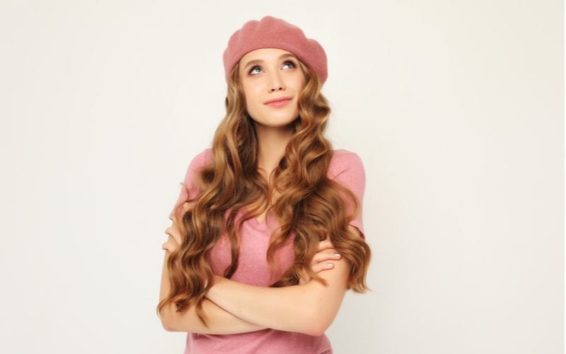 Vrouw met een Frans uitziende hoed met een lange krullende bruine vlecht en een roze shirt kijkt omhoog terwijl ze haar armen kruist.