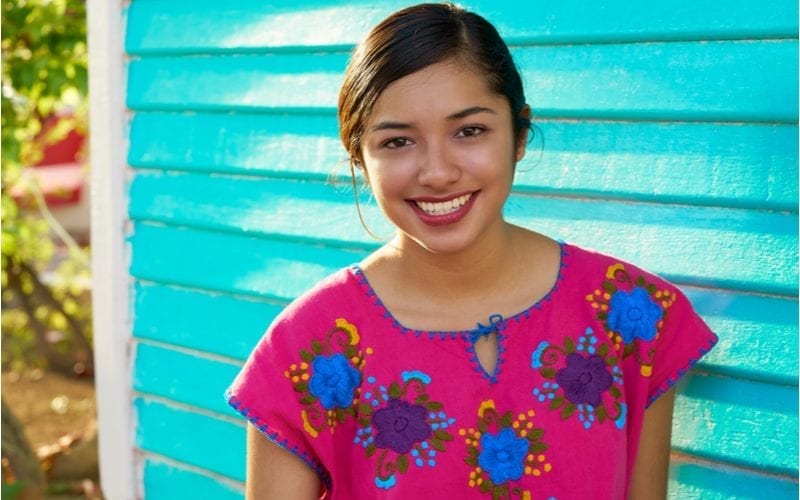 Jonge Mexicaanse vrouw in een roze gebloemd shirt staat voor een verouderd gebouw met blauwe zijkanten.