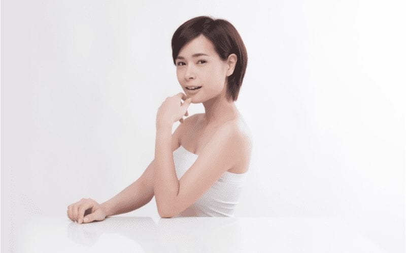 Aziatische kortharige vrouw houdt haar kin vast en zit aan een volledig wit bureau tegen een witte achtergrond