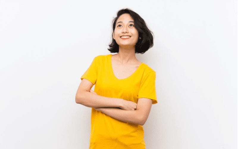 Aziatische vrouw met kort haar en een geel shirt kruist haar armen en kijkt glimlachend omhoog
