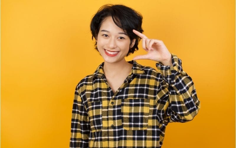 Aziatische vrouw met kort haar in een geel en zwart flanellen hemd houdt haar hand op en glimlacht.