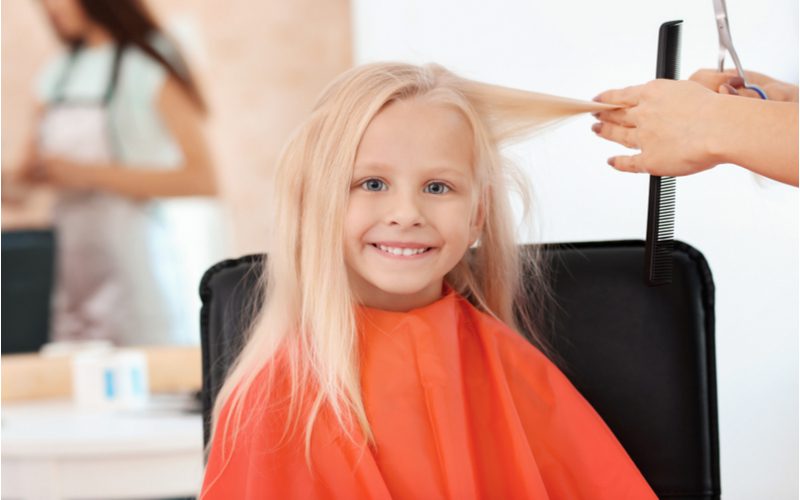 Afbeelding voor een stuk over kleine meisjeskapsels met een jong meisje in een rode cape die haar blonde haar laat knippen terwijl ze glimlacht in een salonstoel.