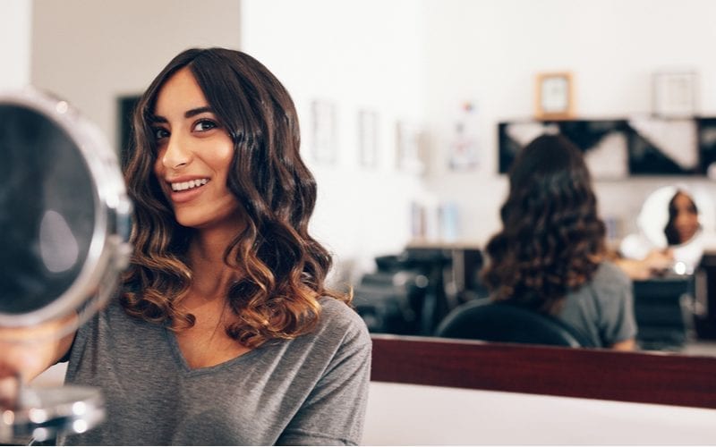 Glimlachende jonge vrouw kijkt naar een handspiegel na de hairstyling. Vrouw kijkt naar de achterkant van haar kapsel in grote spiegel met behulp van een kleine handspiegel in de salon.