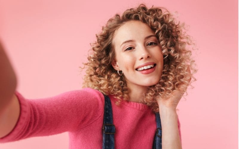 Portret van gelukkig jong meisje met krullend haar dat een selfie neemt die over roze achtergrond wordt geïsoleerd