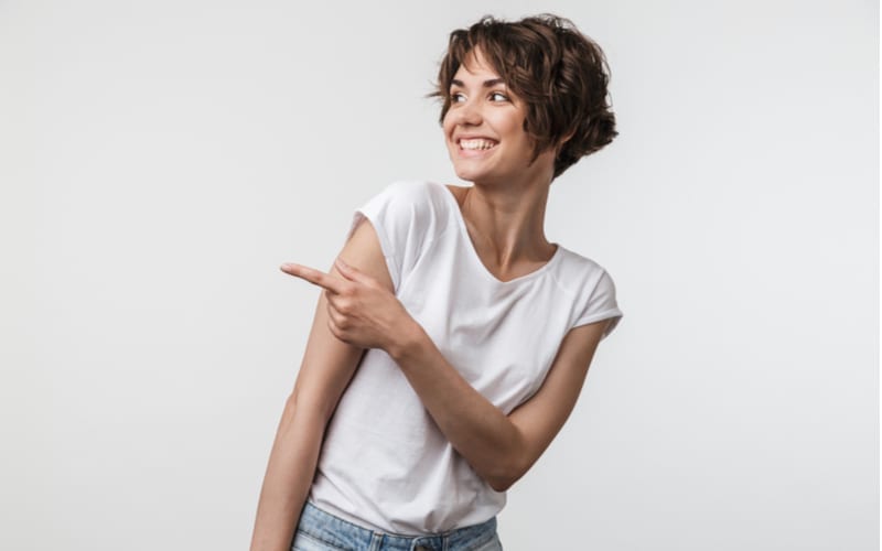 Portret van gelukkige vrouw met kort shaghaar in basis t-shirt dat zich verheugt en vinger op copyspace wijst die over witte achtergrond wordt geïsoleerd