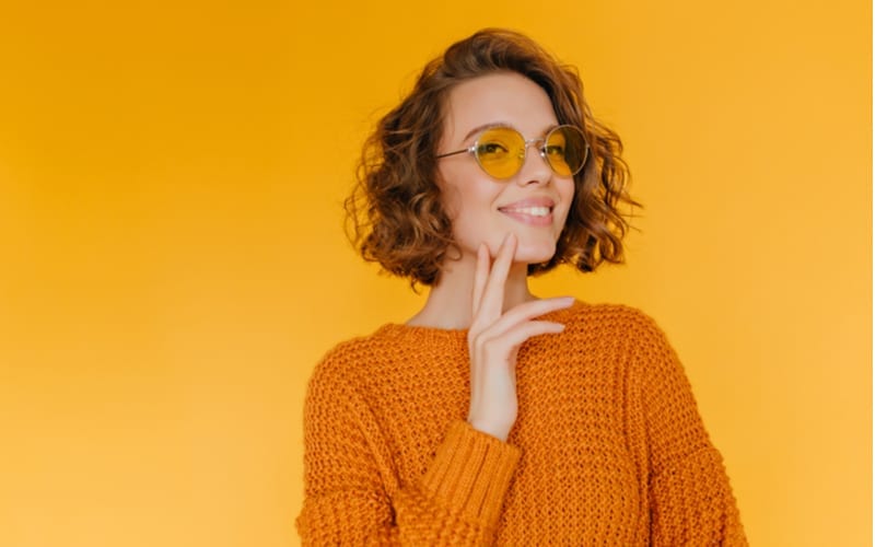 Voor een stuk over korte shagkapsels, Vrolijk kortharig meisje in trendy bril poseert met plezier in nieuwe gebreide kleding. Binnenportret van tevreden krullende jonge vrouw staand voor gele muur.