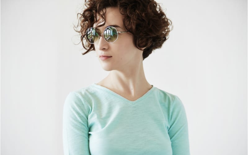 Portret van jonge mooie vrouw met kort krullend shag kapsel die een ronde hipster zonnebril en mint topje draagt, wegkijkend, poserend tegen witte studio muur achtergrond