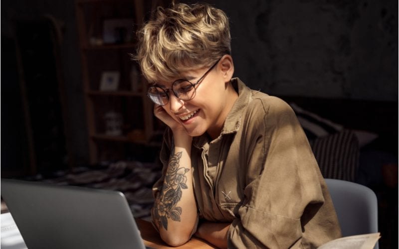 Jonge vrouw kort haar die een bril draagt zit aan bureau die online op laptop afstandsonderwijs bestudeert kijkend naar het scherm lachend vrolijk grappige lezing