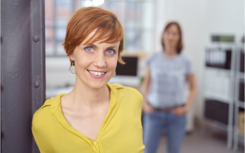 Leuke jonge roodharige vrouw met kort kapsel in gele blouse leunend tegen post in klein kantoor met partner of vriend uit focus op achtergrond