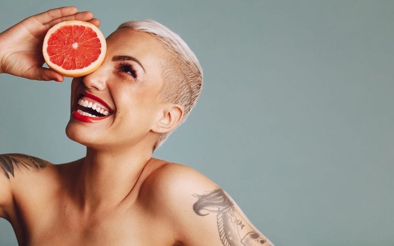 Close-up van een vrouw die een grapefruit voor haar oog houdt tegen grijze achtergrond. Mooi vrouwelijk model met kort blond haar dat een grapefruit schijfje vasthoudt en glimlacht en een Pixie Cut rockt.
