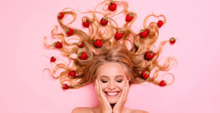 Vrouw met aardbei blond haar letterlijk met het fruit in haar haar liggend op een roze achtergrond en met haar lachende gezicht