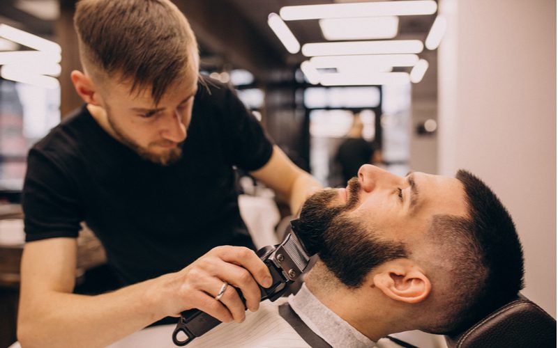 Man met een lage kaalheid krijgt zijn baard getrimd door een kapper terwijl hij achterover ligt in een kappersstoel.
