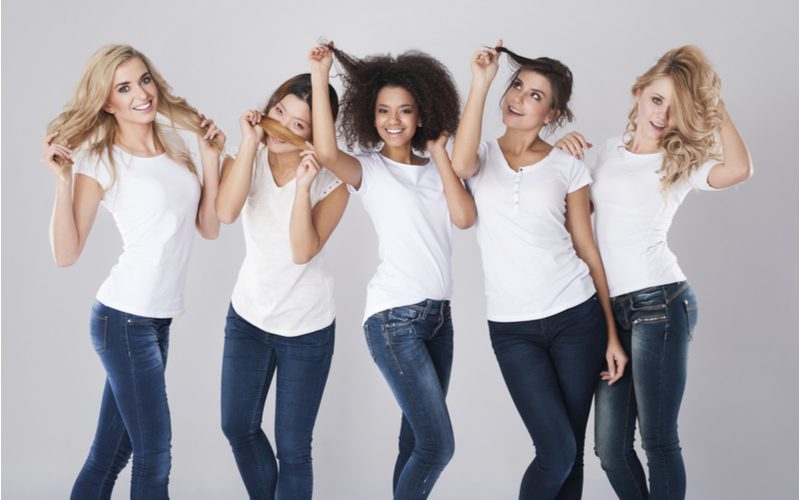 Vrouwen van verschillende rassen en huidskleuren met alle verschillende haartypes uit onze haartype tabel staan in een roze kamer met donkere jeans en witte shirts