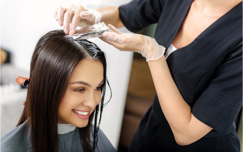 Afbeelding getiteld What Happens If You Leave Hair Dye on Too Long met een vrouw in een salonstoel die glimlacht en highlights krijgt.