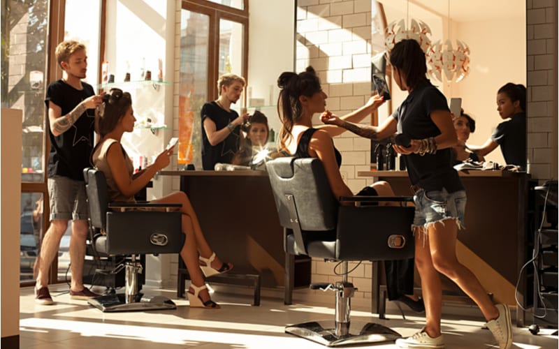 Als beeld voor een stuk over wat is een kapsalon, Beeld van werkdag binnen de schoonheid-zit op twee stoelen klanten mooie jonge meisjes. De kapper maakt haar het stileren of haarknipsel, visagist die make-up in een schoonheidssalon doet-voorraadfoto.