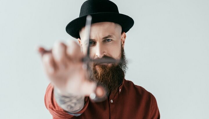 Getatoeëerde en bebaarde barbier met een hoed die een schaar vasthoudt