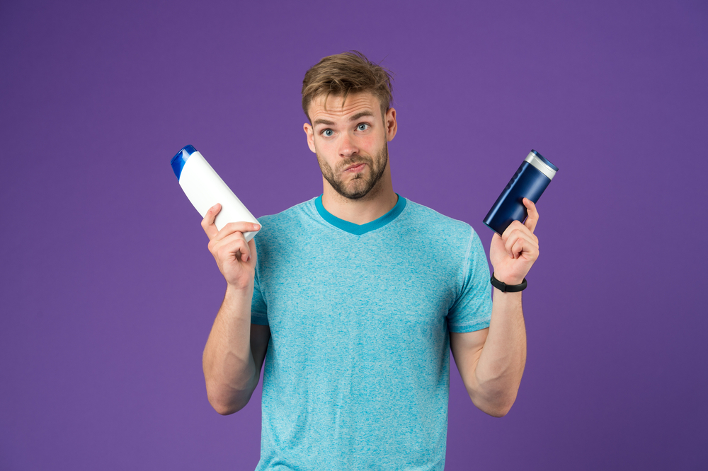 Man houdt twee flessen shampoo voor folliculitis vast terwijl hij een veeggezicht maakt met zijn lippen