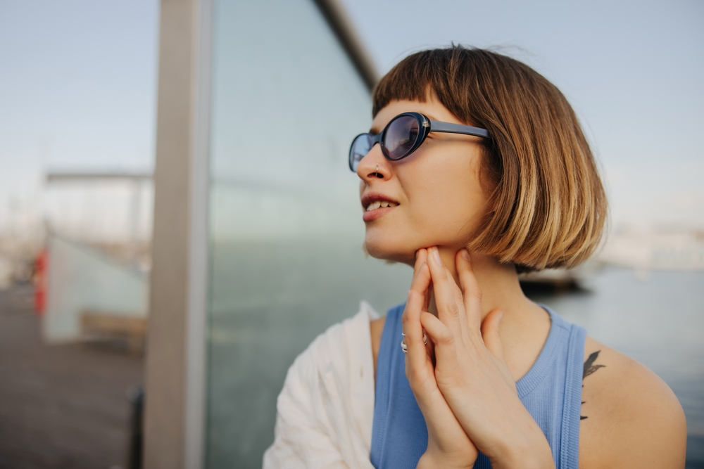 Vrouw met somber kort haar en blauwe zonnebril kijkt weg in de verte met de handen ineengeslagen