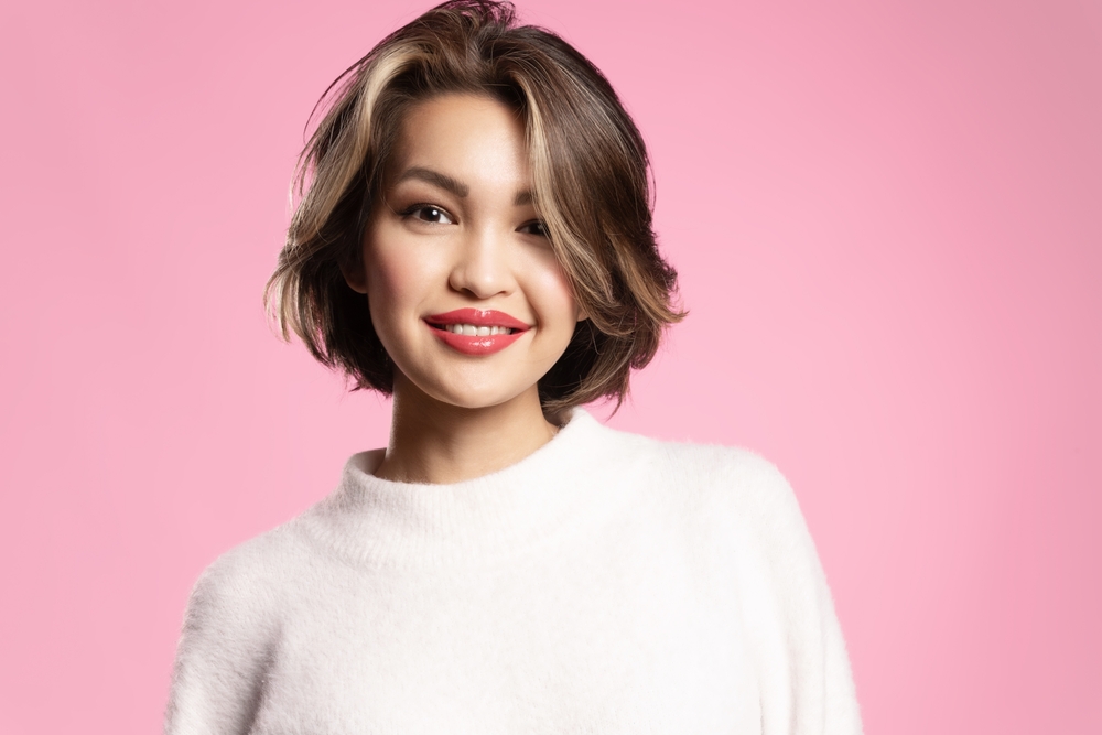 Vrouw glimlacht naar camera met haar over één oog op roze achtergrond om de beste haarkleur voor kort haar te tonen