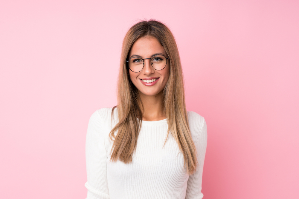 Glimlachende vrouw met bril staat voor roze muur met donker goudblond haar om de beste haarkleur voor leeftijdsverandering te laten zien