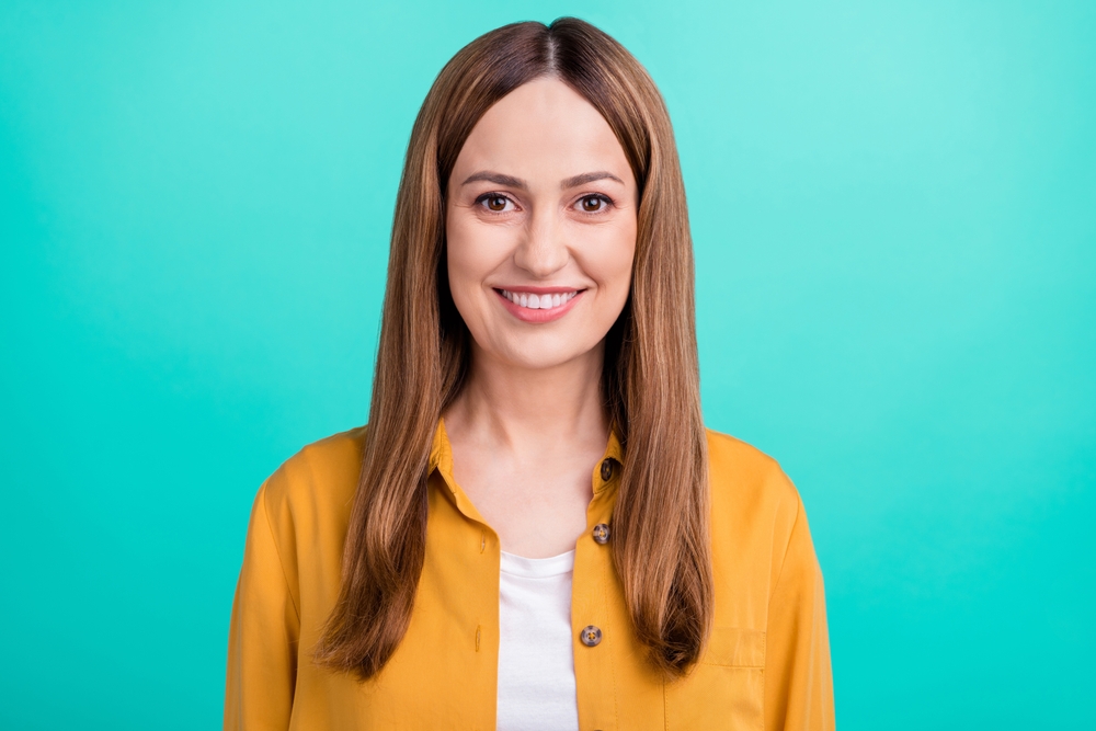 Vrouw in de 40 lacht voor turquoise muur met geel vest en draagt een van de beste haarkleuren voor leeftijdswisseling in brons