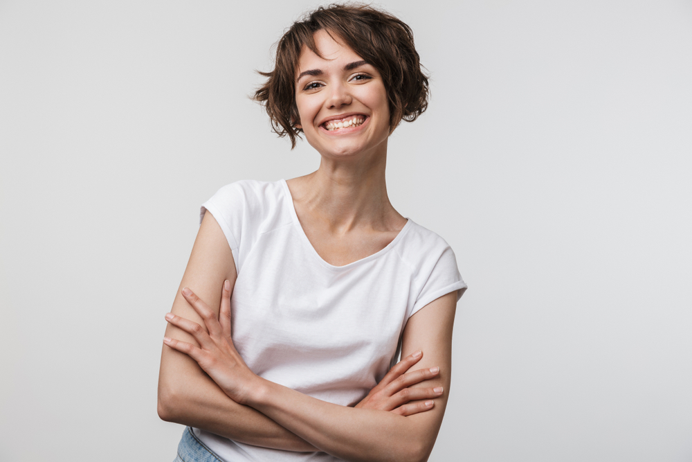 Glimlachende brunette vrouw met wit t-shirt tegen witte achtergrond kruist haar armen met een korte bob en zeer piekerige pony's