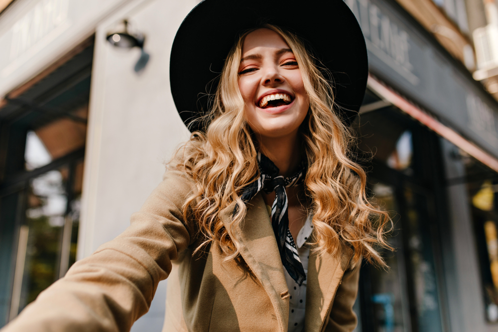 Jonge vrouw met krullend honingblond haar draagt trendy hoed en jas glimlachend in de stad