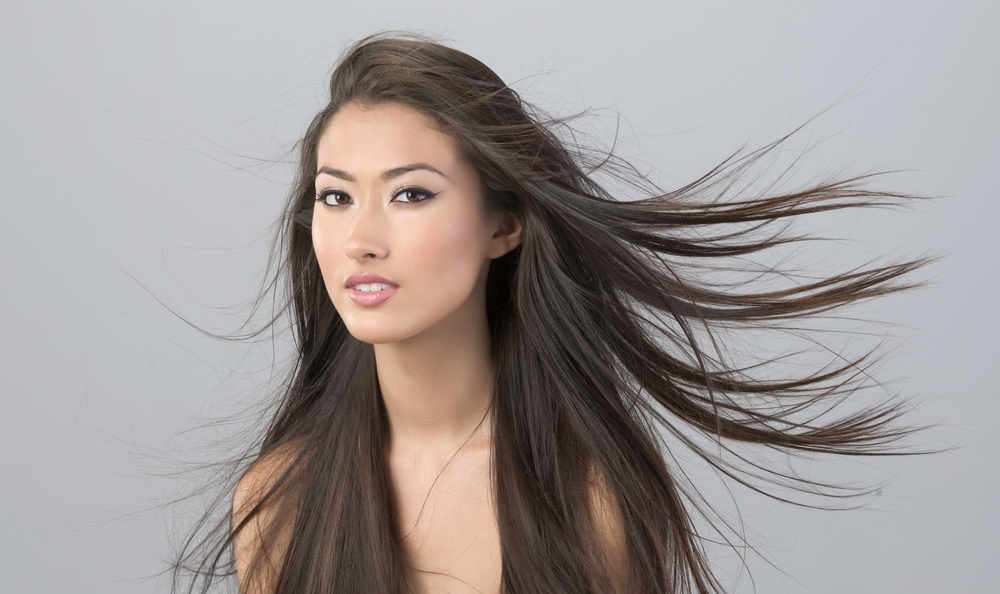 Aziatische vrouw kijkt naar de camera met haar as-getinte donkerbruine haar wapperend in de wind voor een grijze muur.