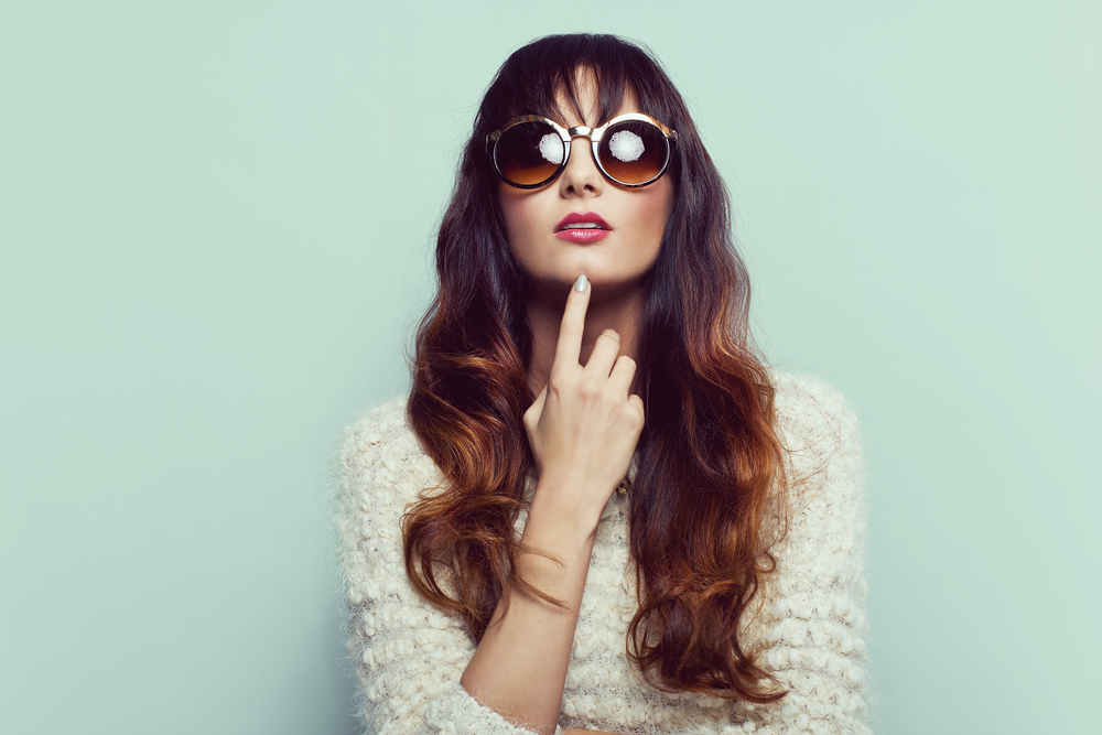 Vrouw plaatst vinger op kin en draagt grote ronde zonnebril voor mintkleurige muur met lang donkerbruin haar en auburn ombre