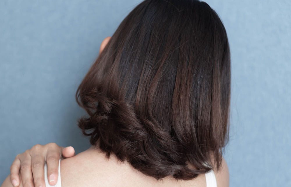 Achteraanzicht van vrouw met hand op haar schouder die pronkt met een lange bob met gekrulde uiteinden en donkerbruine kleur