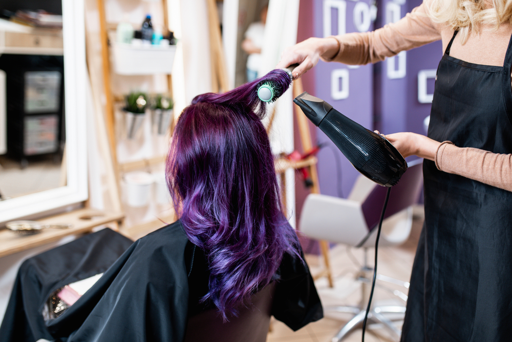 Hairstylist droogt het pas geverfde donkerpaarse haar van de klant met een ronde borstel in een moderne kapsalon