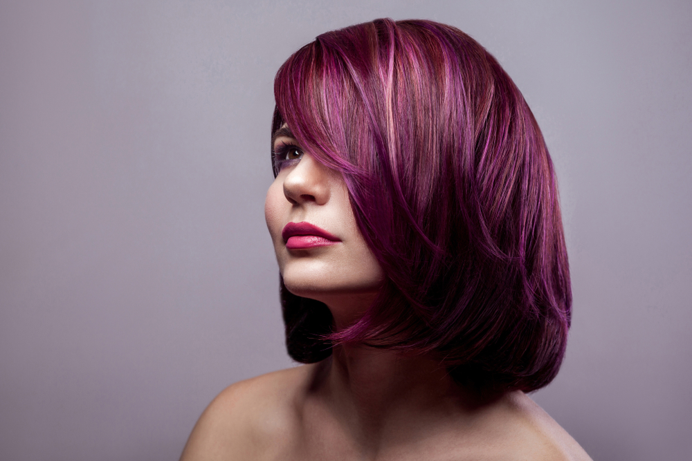 Vrouw kijkt opzij met blote schouders met een donkerroze paarse haarkleur voor een grijze muur