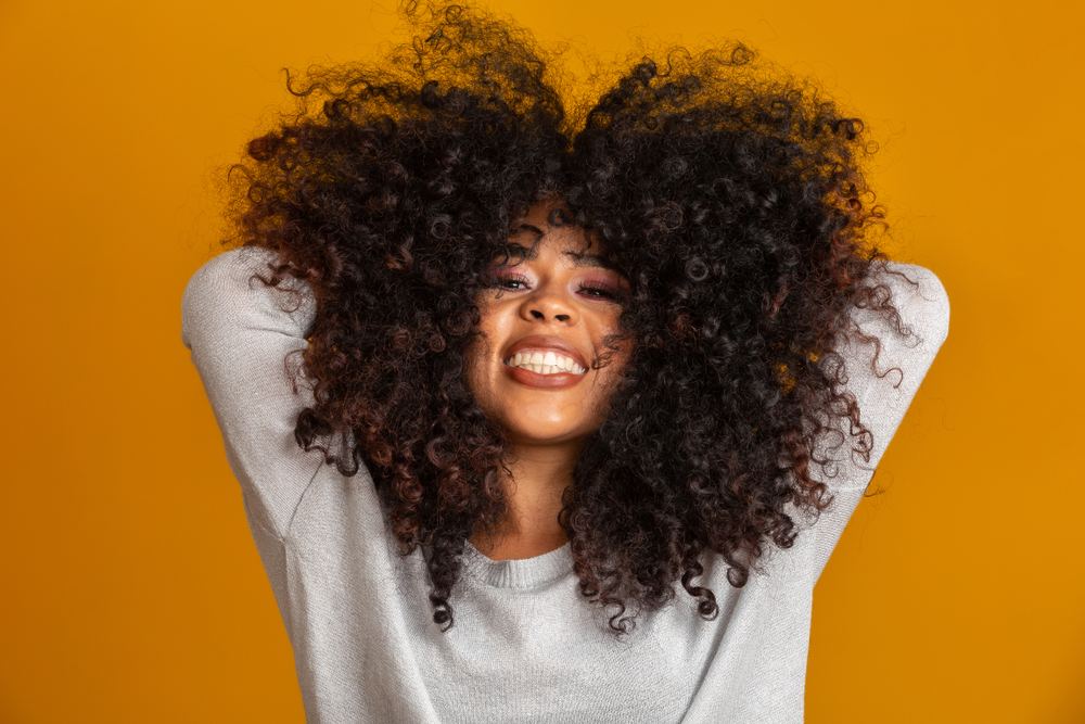 Zwarte vrouw houdt haar handen achter haar hoofd voor de oranje muur en toont haar gezonde haartextuur die kan worden bereikt met behulp van black owned haarproducten.