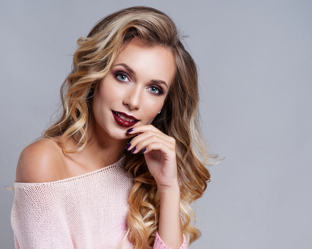 Blonde vrouw poseert met haar hand in de buurt van haar mond met een off shoulder roze top en glam krullen voor lang dun haar.