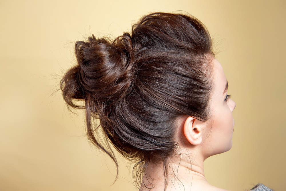 Achteraanzicht van vrouw voor gele muur met haar dunne lange haar in een rommelig knotkapsel met extra volume vooraan