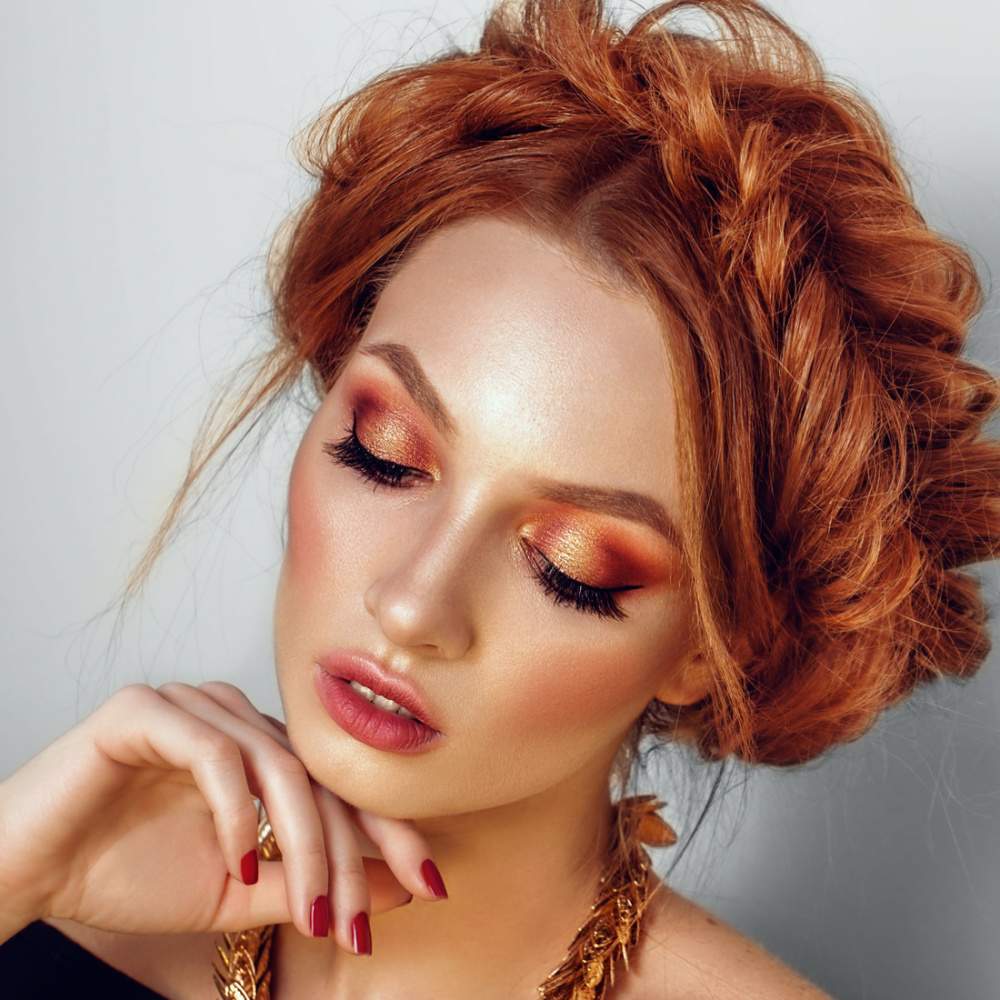 Roodharige vrouw met koperen glam make-up look heeft haar haar gestyled in een fishtail kroonvlecht met hand geposeerd onder haar kin.