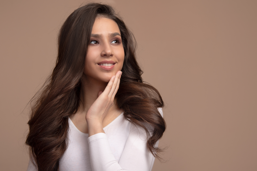 Brunette vrouw met lang dun haar gestyled in een volumineuze blowout poseert met haar hand op haar kin voor een lichtbruine achtergrond in een wit v-hals shirt.