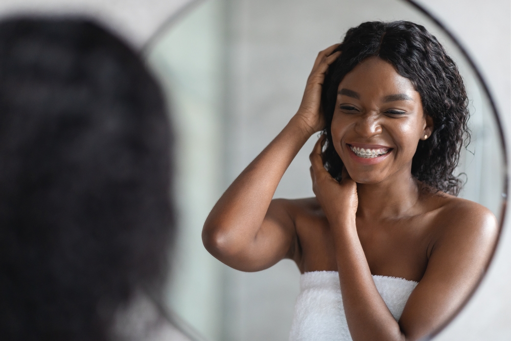 Zwarte vrouw glimlacht in spiegel terwijl ze haar pas gewassen haar styled met producten gemaakt door zwarte bedrijven.