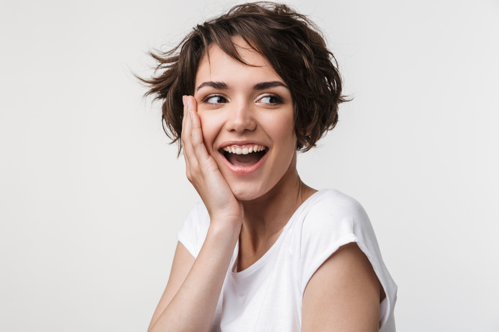 Brunette vrouw met bob en pony stijl houdt haar hand tegen haar wang in verbazing met een glimlach en witte shirt op