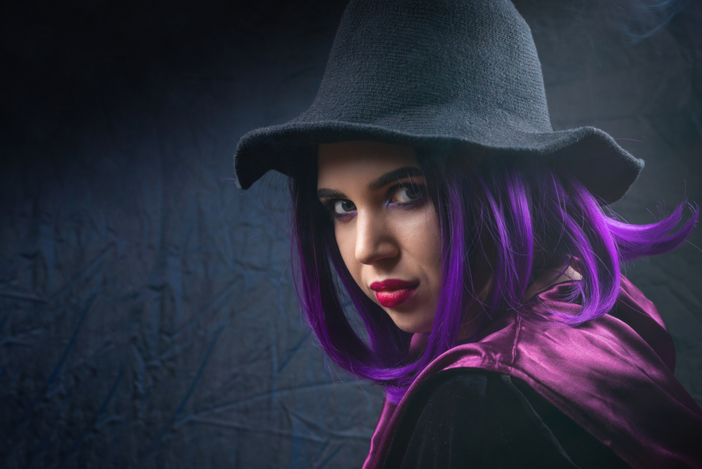 Vrouw met paars haar verkleed als heks voor Halloween draagt zwarte hoed en kijkt over haar schouder