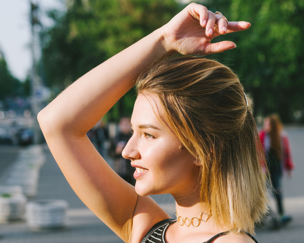 Vrouw met blond balayage haar draagt één van de gemakkelijkste half op half neer kapsels met gezichtsbepalende lokken en arm over haar hoofd geheven.