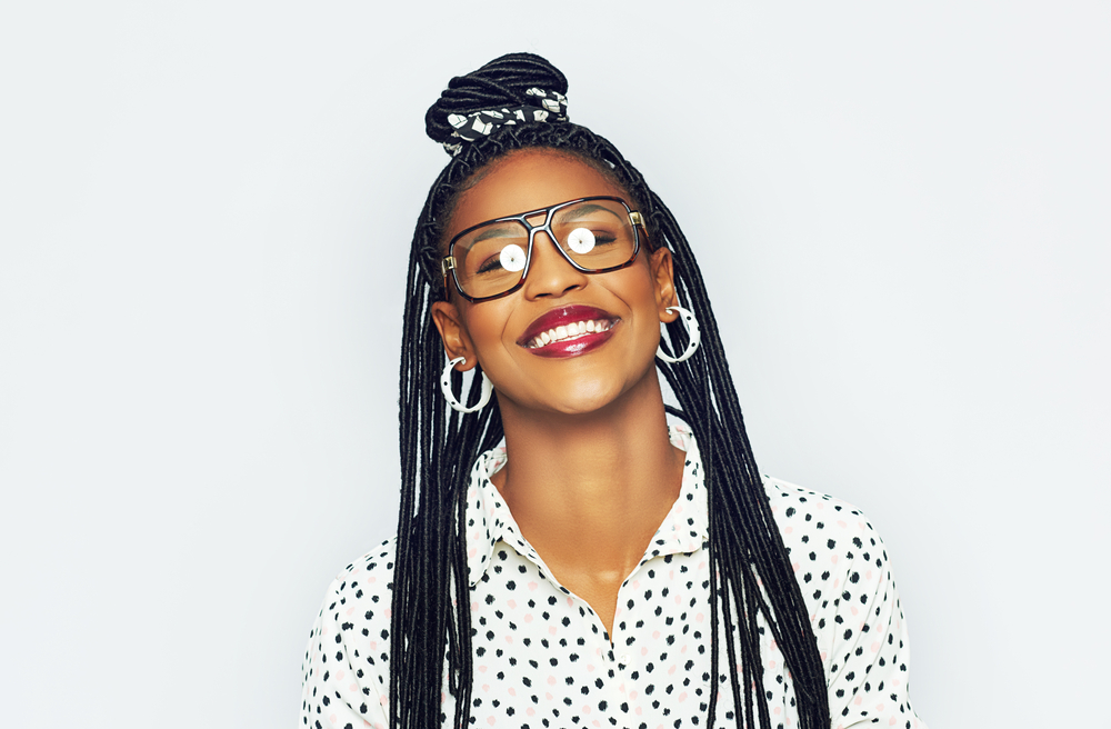 Afro-Amerikaanse vrouw met bril en witte top met stippen draagt oorbellen en half op half neer knot stijl voor witte achtergrond.