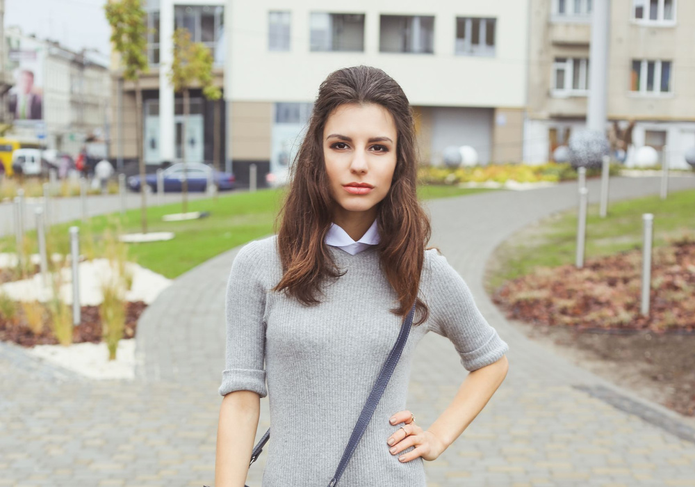 Brunette vrouw in grijze gebreide jurk met crossbody tas modelleert één van haar half op half neer kapsels buiten