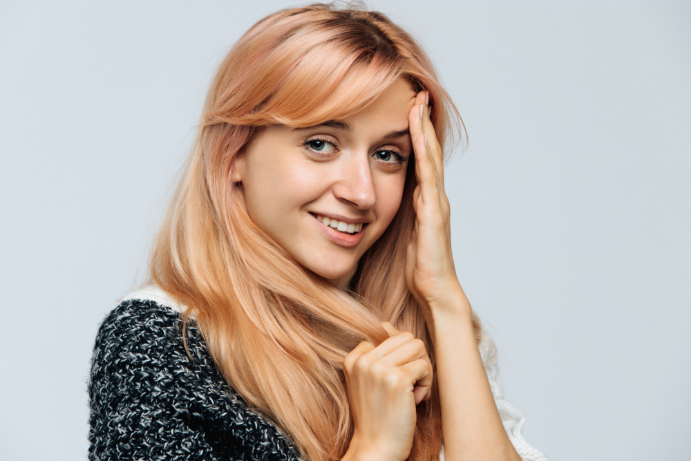 Vrouw glimlacht met hand die haar voorhoofd aanraakt om één van de unieke tinten blond te tonen in een rosé gouden kleur met lang haar en pony aan de zijkant.