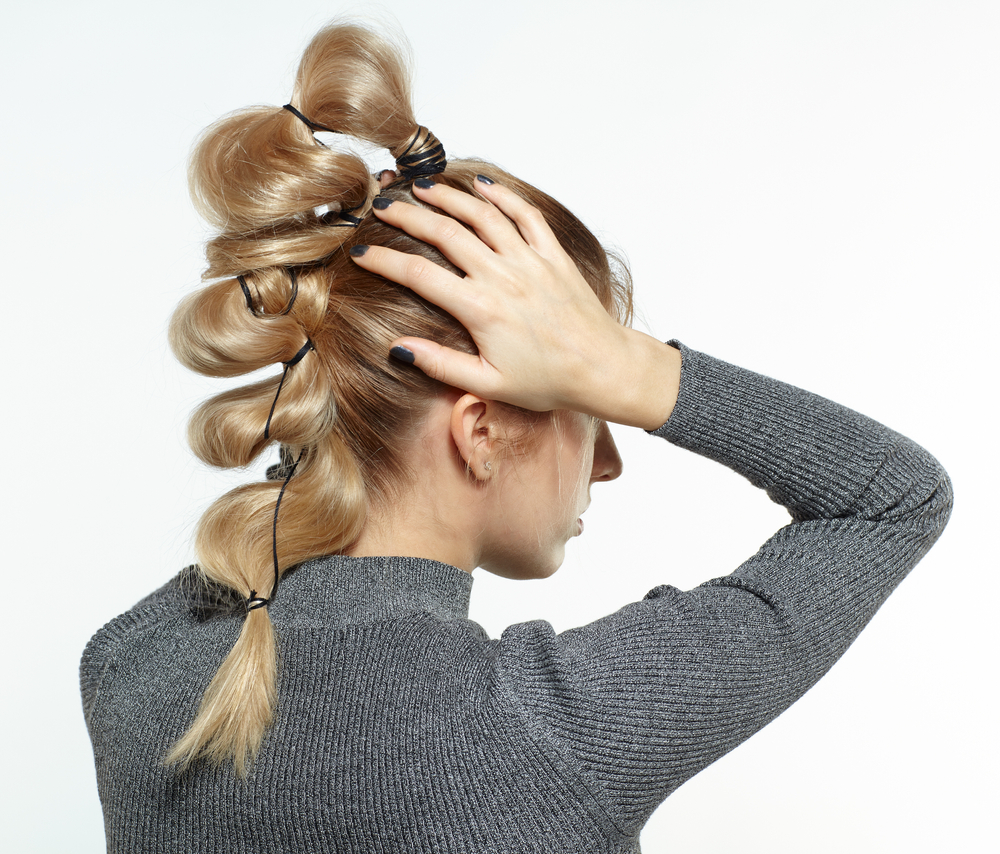 Achteraanzicht van blonde vrouw met handen op haar hoofd die een looped bubble braid kapsel draagt voor witte muur